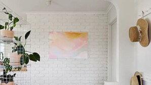 A room with a white brick wall, a wooden floor, a pink abstract painting, and a shoe rack with hats. - Peachbud House Portarlington (Portarlington)
