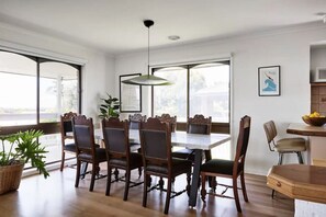 A dining area with a large wooden table and chairs, a hanging light fixture, and large windows with a view.