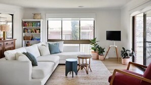 A modern living room with a white sofa, a small round table, and a red chair. There is a bookshelf filled with books and a television mounted on the wall. - Peachbud House Portarlington (Portarlington)