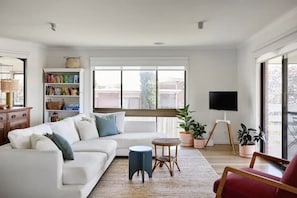 A modern living room with a white sofa, a small round table, and a red chair. There is a bookshelf filled with books and a television mounted on the wall.