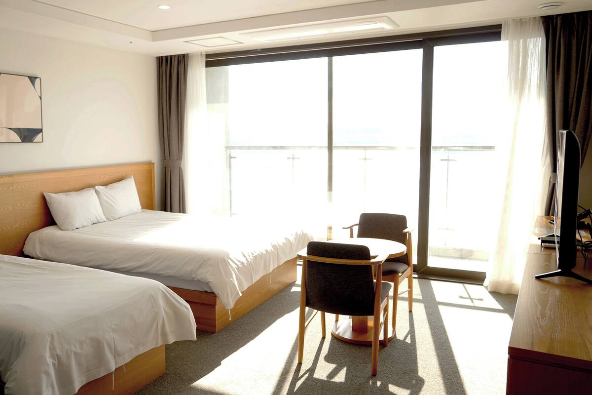 A hotel room with two beds, a small table with a chair, a TV, and a large window with curtains.