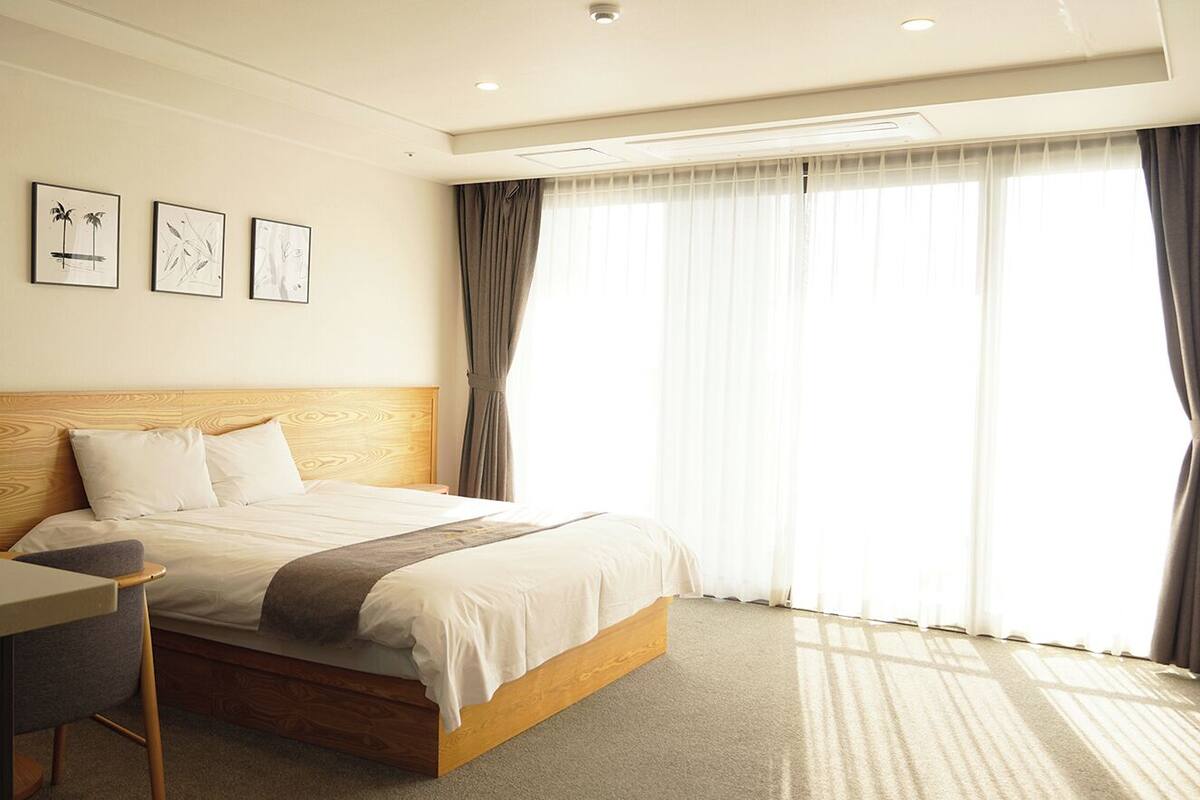 A hotel room with a wooden headboard, a bed with white bedding, a desk, and a large window with curtains.