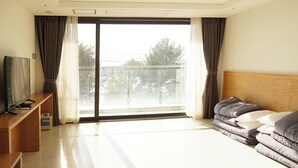 A hotel room with a large sliding glass door to a balcony, a bed with grey bedding, a wooden headboard, a TV on a wooden stand, and a small table with a remote control. - Hotel Ocean Hill (Yeosu)