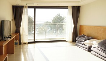 Habitación de hotel con un amplio ventanal corredera a un balcón, una cama con ropa de cama gris, un cabecero de madera, un televisor sobre un mueble de madera y una mesita con un control remoto.