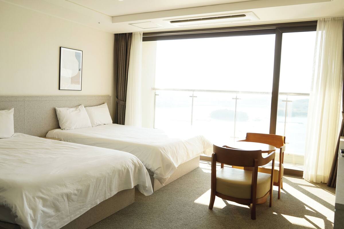 A hotel room with two beds, a small table, and a chair. There is a large window with curtains and a view of the outside.