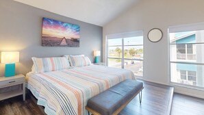 Ein Schlafzimmer mit einem großen Bett, einer Bank, einem Nachttisch, einer Lampe, einem Sonnenuntergangsbild und Blick ins Freie.