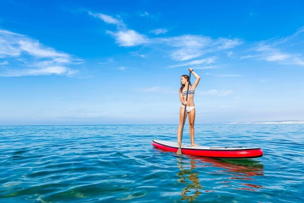 A person standing on a red paddleboard in clear blue water with a distant shoreline. - Resort Style Community. Quick Walk to Beach. Heated Pool. Near Great Restaurants & Shops. (Madeira Beach)