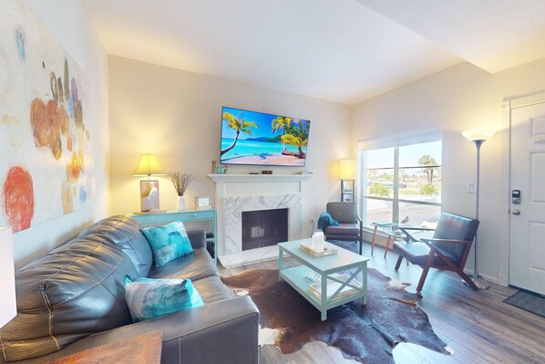 A living room with a sofa, coffee table, and chairs, a fireplace, and a TV displaying a tropical scene.