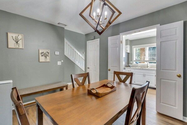 A dining area with a wooden table, chairs, and a chandelier. There are framed pictures on the wall and a staircase leading to another room.