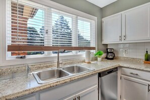 A kitchen with a double sink, white cabinets, a window with blinds, and a coffee maker.