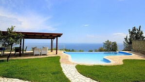 A swimming pool with a clear view of the sea, a covered seating area, and a stone pathway.