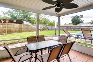 Eine überdachte Terrasse mit Tisch und Stühlen, einem Deckenventilator und Blick auf einen Garten mit Pool und Holzzaun.