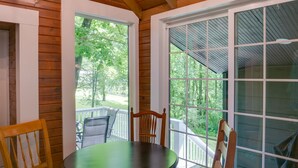 Una terraza de madera con una mesa y sillas, ventanas amplias y vistas a árboles.