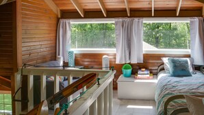 Una cabaña con interior de madera, una cama con almohada azul, una ventana con cortinas y una mesita con libros.
