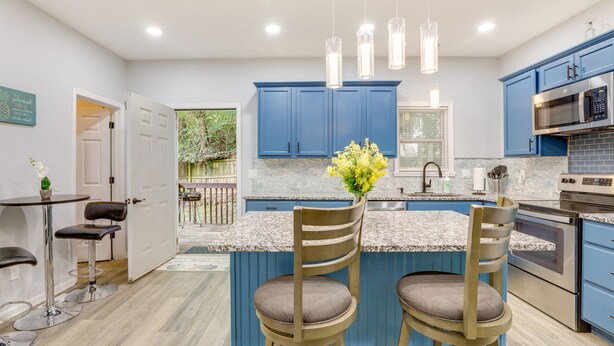 Een moderne keuken met blauwe kastjes, een centraal kookeiland met twee barkrukken, roestvrijstalen apparatuur en hanglampen.