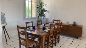 Une salle à manger avec une table en bois, six chaises, une plante en pot et une fenêtre avec des stores.