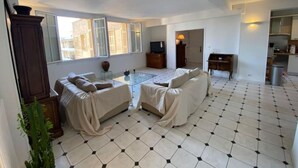 Un salon spacieux comprenant un ensemble de canapés, une table basse et un téléviseur. La pièce est dotée de grandes fenêtres et d’un plancher carrelé.