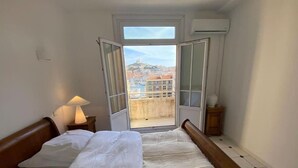 Une chambre à coucher avec un lit, une table de chevet avec une lampe, et une vue sur les bâtiments à travers une porte ouverte.