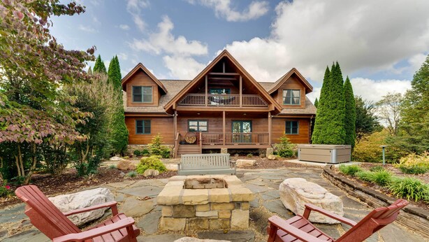 A wooden cabin with a balcony, stone fire pit, and Adirondack chairs.