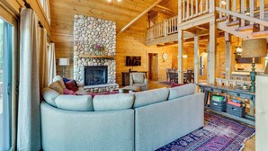 A cozy cabin interior with a stone fireplace, a large sofa, a dining area, and a wooden staircase.