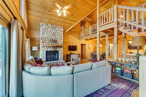 A cozy cabin interior with a stone fireplace, a large sofa, a dining area, and a wooden staircase.