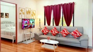 A modern living room with a grey sofa, red and white patterned cushions, a flat-screen TV, a small white table, and a bed with a white headboard.