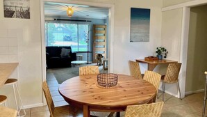 A dining area with a round wooden table, herringbone-patterned chairs, a hanging light fixture, and a view into the living room through an open doorway.