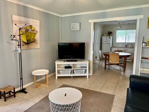 Una sala de estar con televisor, una mesita, un taburete, una lámpara de pie y una cocina al fondo.