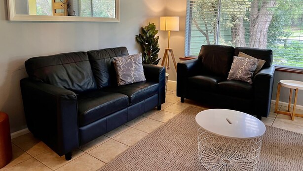 A living room with a black leather sofa, a blue armchair, a white coffee table, and a large window with blinds.