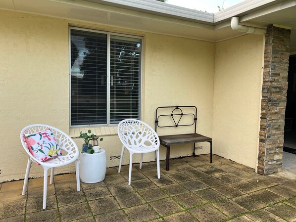 Un patio con dos sillas blancas, una mesa pequeña con una planta y un banco.