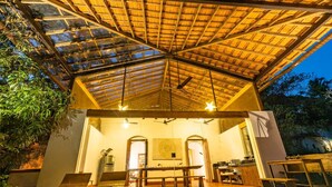 Een openluchtdinerruimte met een hoge plafond en houten balken. Tafels en stoelen staan klaar voor diners, en een keuken is zichtbaar op de achtergrond.