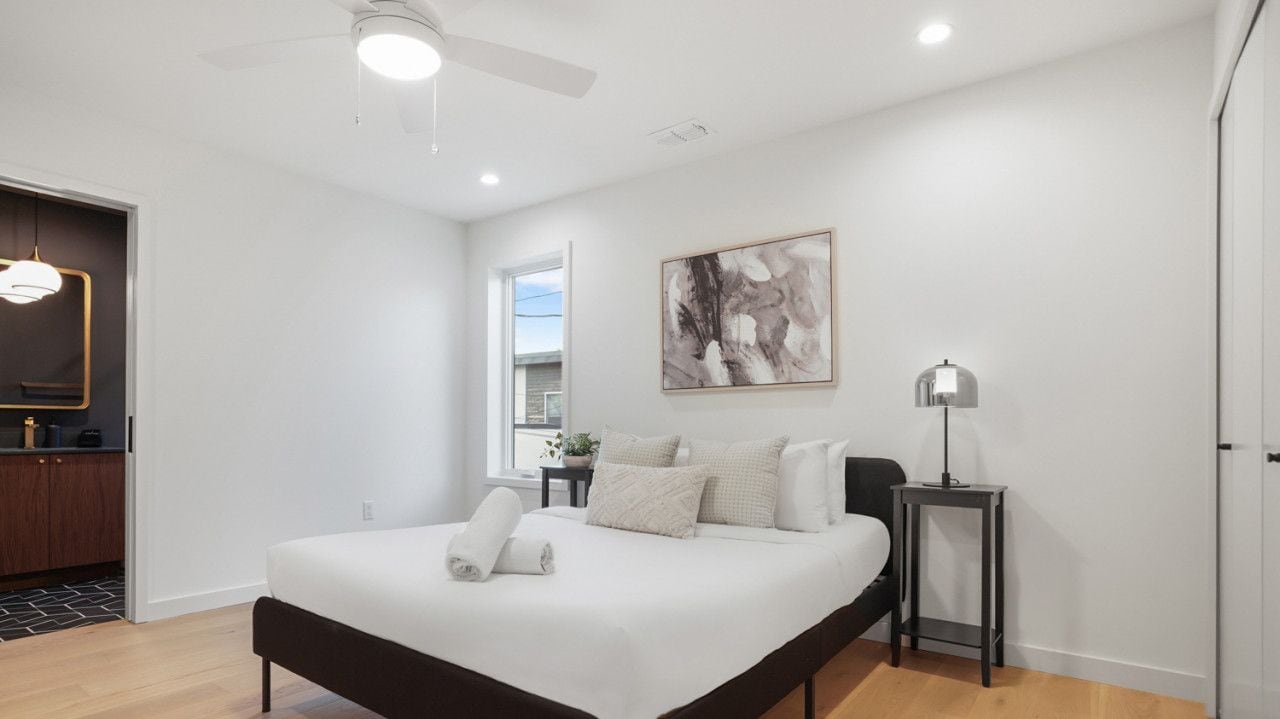 A modern bedroom with a large bed, a ceiling fan, a bedside table with a lamp, and a window with a view.