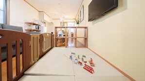 A hotel room with a tatami mat floor, a wooden crib, a television mounted on the wall, and a kitchen area visible through an open door. - HATAGAYA Apartment 1F (Tokyo)