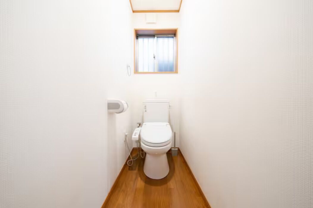 A small, narrow bathroom with a toilet, a window, and a wooden floor.
