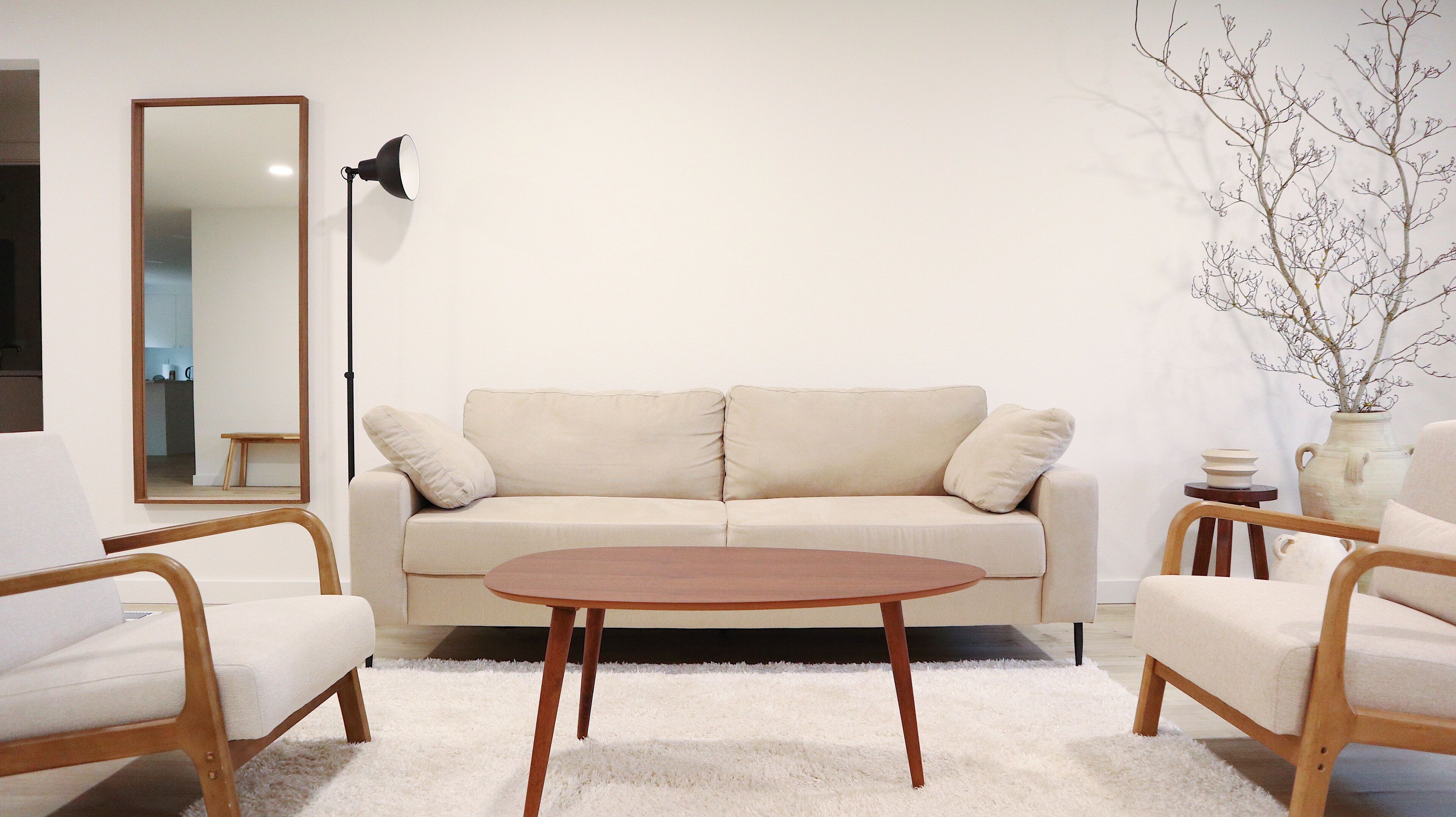 Un salón minimalista con un sofá color beige, dos sillas de madera, una mesa de centro redonda de madera, una lámpara de pie y un jarrón blanco con ramas sobre una mesita auxiliar.