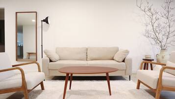 Un salón minimalista con un sofá color beige, dos sillas de madera, una mesa de centro redonda de madera, una lámpara de pie y un jarrón blanco con ramas sobre una mesita auxiliar.