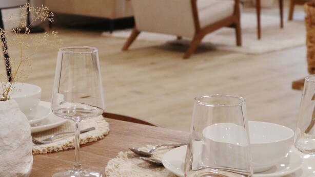 Una mesa de comedor dispuesta con platos blancos, vasos y cubiertos, rodeada de sillas color beige y un sofá en una habitación de tonos neutros.
