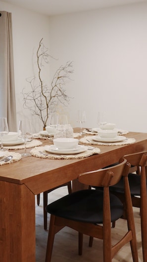 Une table de salle à manger en bois, dressée avec de l’assiette blanche, des bols et des verres à vin, agrémentée d’un vase contenant des branches.