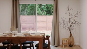 Un comedor con una mesa lista para cuatro personas, una silla de madera, un taburete pequeño y un florero con ramas.