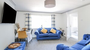 A living room with a blue sofa, a dining table, and a TV mounted on the wall. - 22 Turnberry Road (Girvan)
