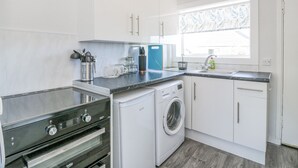 A modern kitchen with white cabinets, a built-in oven, a washing machine, and a window with a leaf-patterned blind. - 22 Turnberry Road (Girvan)