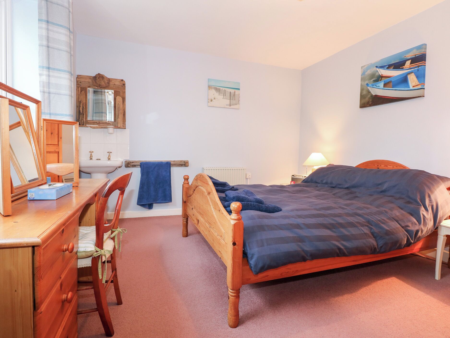 A bedroom with a wooden bed, a desk with a chair, a mirror, a sink, and a towel rack.