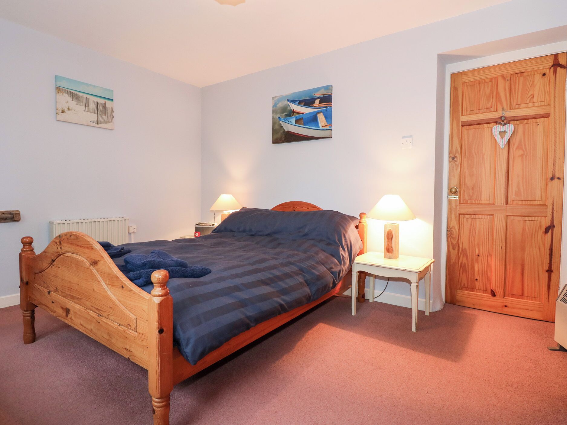 A bedroom with a wooden bed, two bedside tables with lamps, a wooden door, and two wall-mounted paintings.