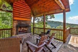 A wooden deck with a fireplace, wicker furniture, and a view of a forested hill.