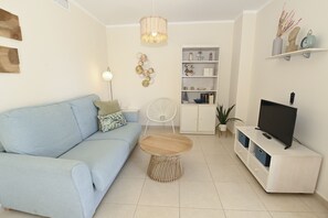 Uma sala de estar com um sofá azul-claro, uma mesa de centro redonda, um ventilador branco, uma TV sobre um móvel branco e arte decorativa na parede.