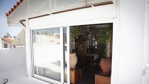 Un edificio blanco con un amplio ventanal corredera que da a un espacio exterior con plantas en macetas y una mesa de madera.