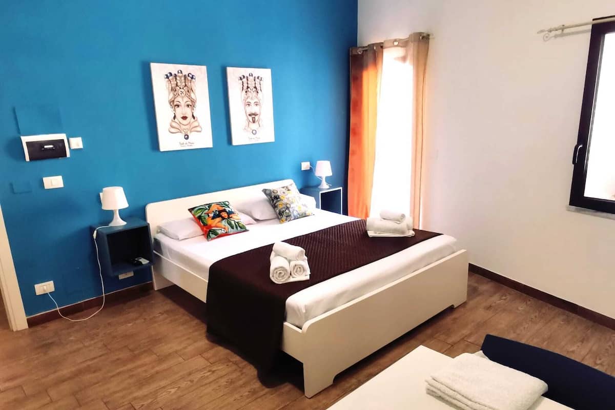 Una habitación de hotel con cama, mesitas de noche, cortinas en la ventana y cuadros en la pared.
