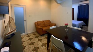A compact room with a sofa, a dining table with a vase of flowers, a small sailboat model, and a bed with a blue curtain. - Historic Ocean Park Near Beach Water Reserve Fiber Internet Fast WiFi (San Juan)
