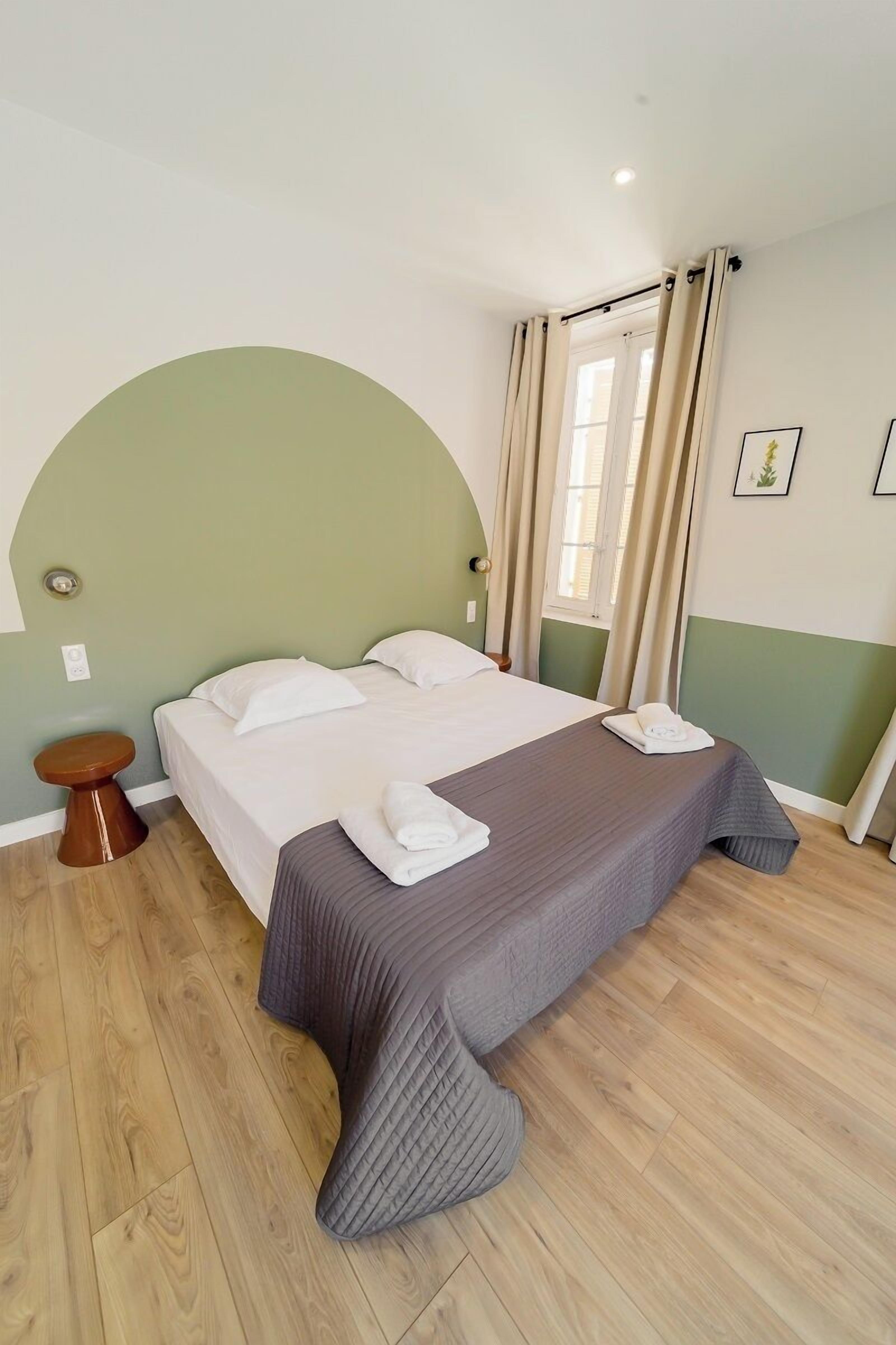 Un lit bien fait, avec une couvre-lit grise et des oreillers blancs, une table de chevet en bois et un petit tabouret rond.