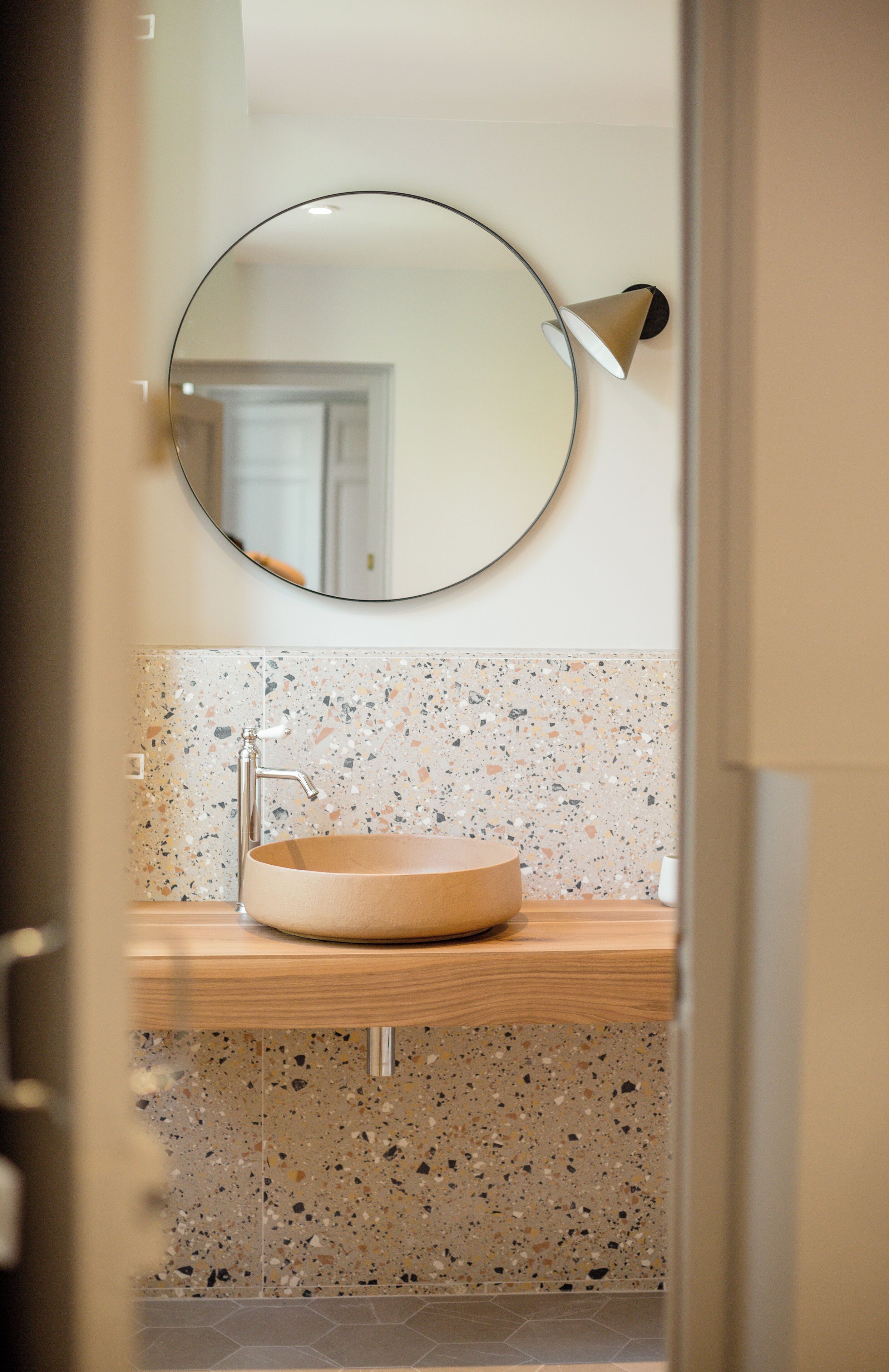 A bathroom with a round mirror, a wooden vanity, and a speckled tile backsplash.